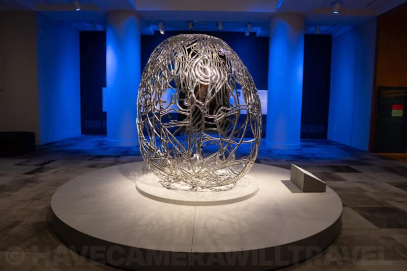 Silvery wire-sculpted egg-shaped artwork glowing under blue gallery lighting