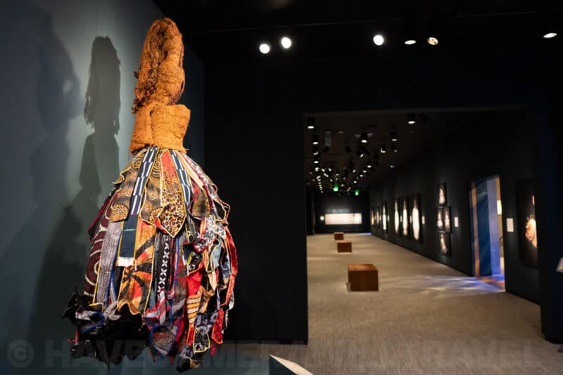 Handmade dress of woven fabric strips hanging beside a dim museum hallway