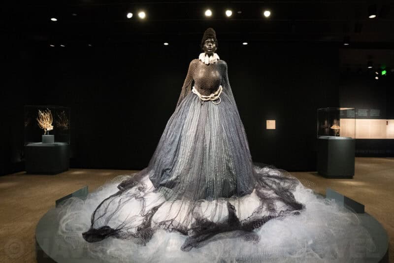 Sculpted figure in a flowing metallic mesh gown displayed in a dark gallery