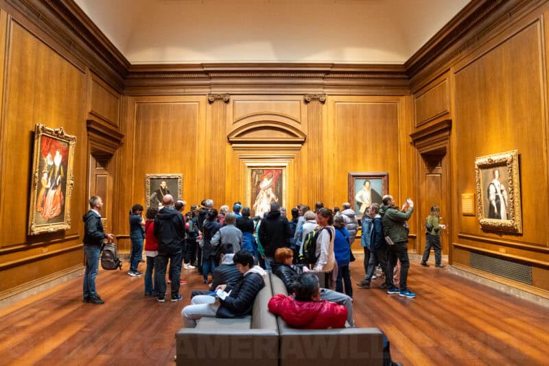 Museum visitors gathered to view framed portrait paintings in a wood-paneled gallery