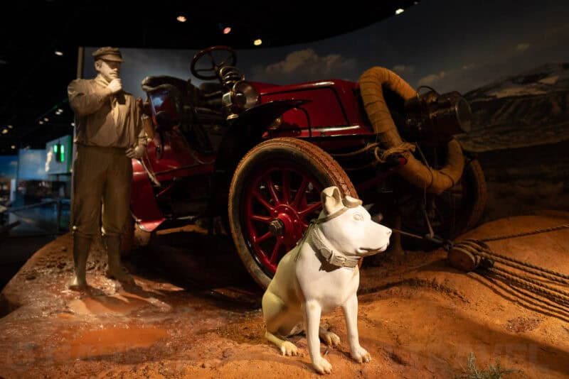 Museum diorama of a vintage red car with a driver figure and a seated dog statue