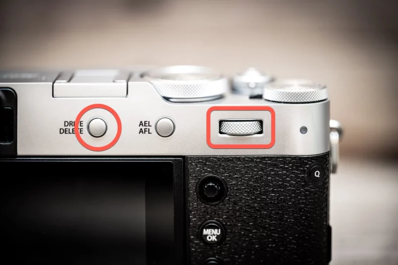 Top of a silver Fujifilm X100VI with the Drive/Delete button and rear command dial circled in red