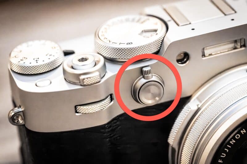 Close-up of the Fujifilm X100VI viewfinder selector lever on the front of the silver camera body, circled in red
