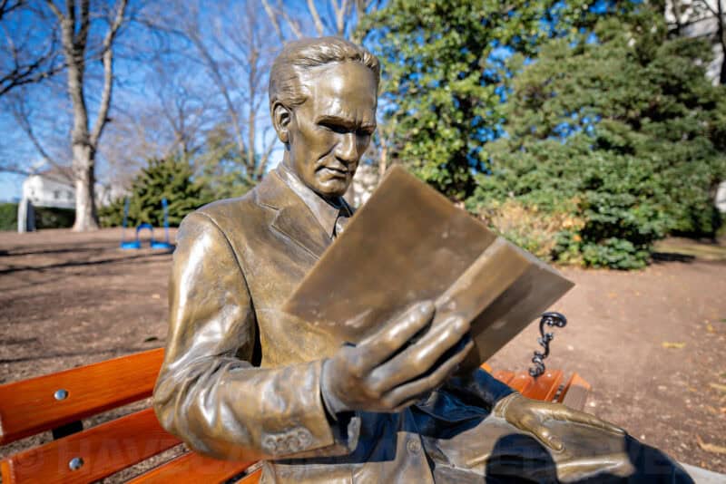 Bronze statue of a seated man reading a book on a park bench outdoors