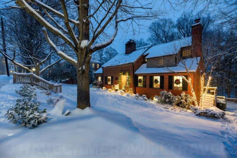 Snow-covered brick colonial house lit warmly with holiday wreaths and lights at dusk