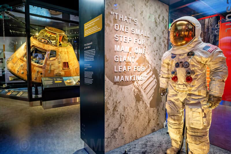 Apollo command module and Neil Armstrong spacesuit display beside a moon-landing quote wall in a museum