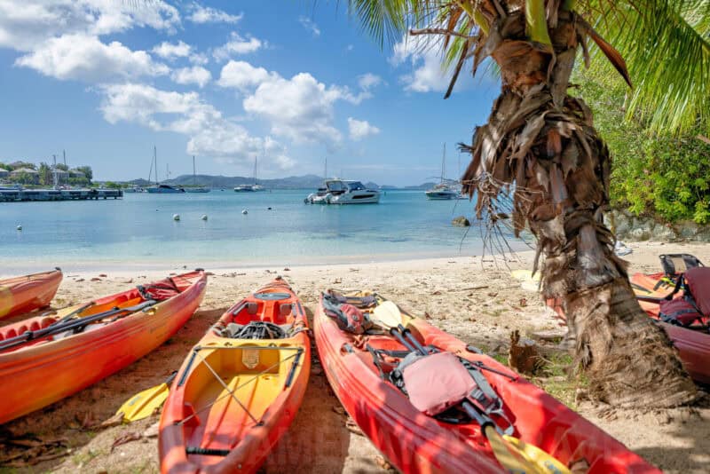 Red kayaks lined up on a sandy beach beside a palm tree with boats anchored in turquoise water