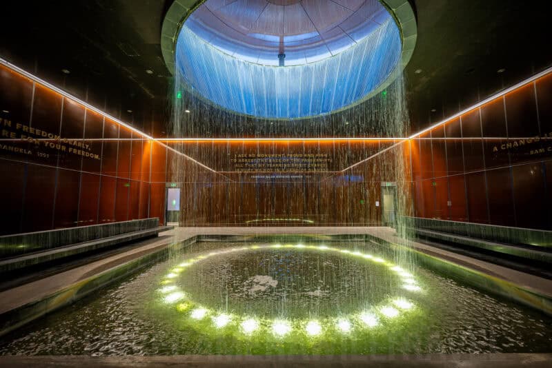 Illuminated water feature pouring through a circular skylight into a reflecting pool at a memorial