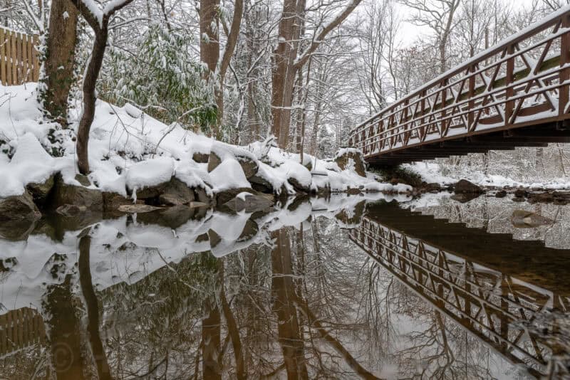 Snow-covered rocks and trees beside a wooden footbridge reflected in a calm stream