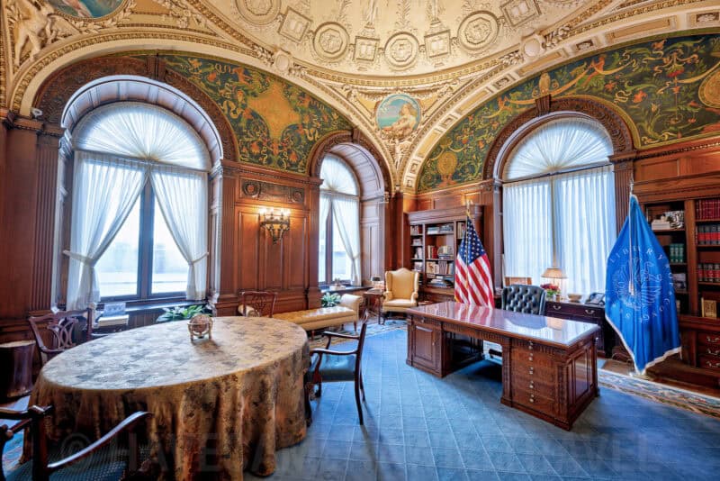 Wood-paneled office with arched windows, mural ceiling, desk and American flag