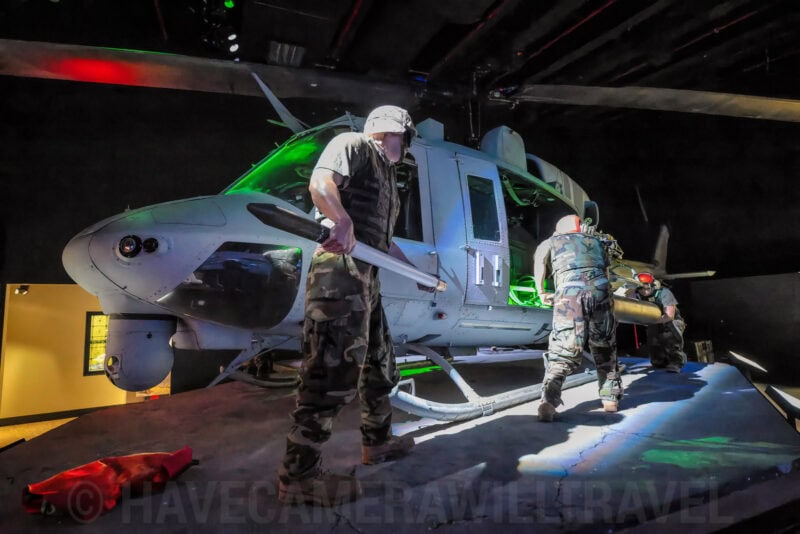 Soldiers in camouflage examining a mounted military helicopter under green stage lighting