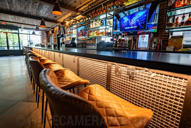 Long leather-topped bar with rows of stools facing shelves of liquor bottles and TV screens