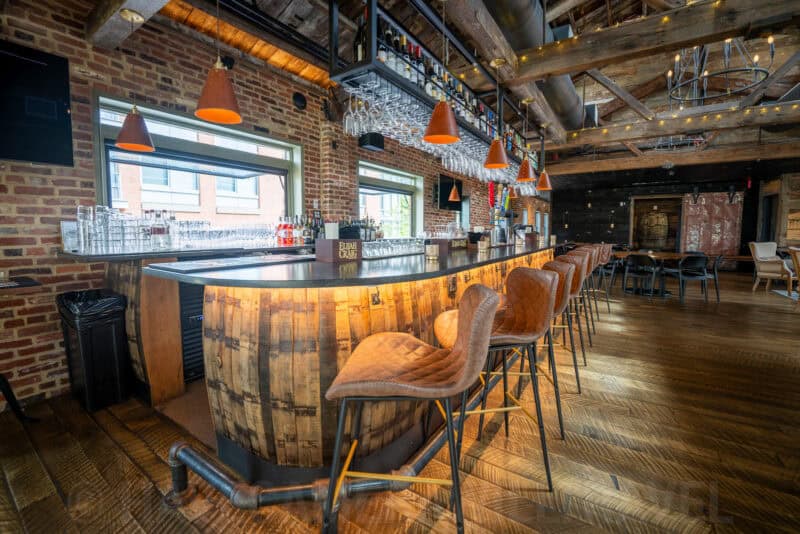 Whiskey-barrel bar counter with leather stools under hanging wine glasses and string lights
