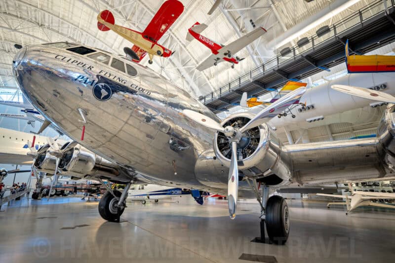 Polished silver vintage propeller airliner nose on display beneath small aircraft in a museum hangar