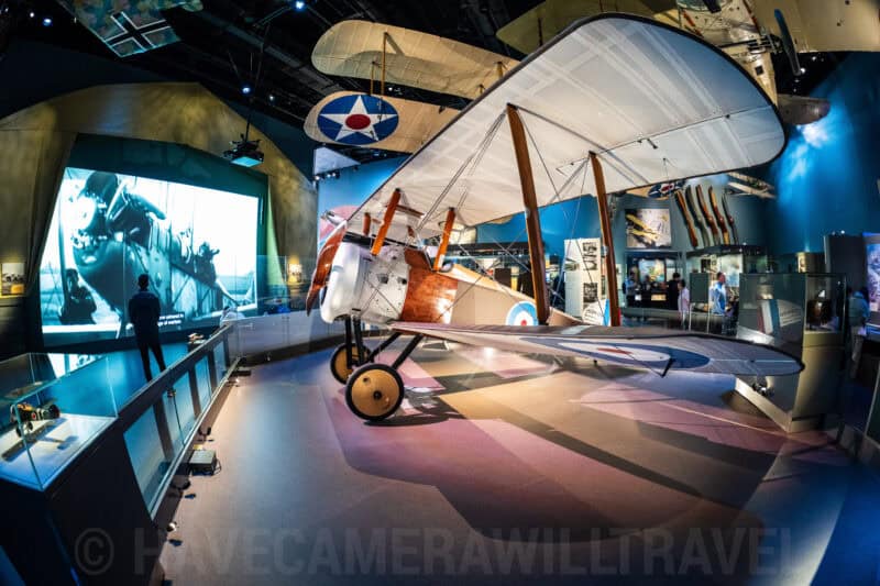 Vintage biplane on display inside a museum gallery with a large screen showing archival war footage