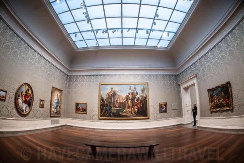 Fisheye view of a skylit gallery room with large framed paintings on patterned walls