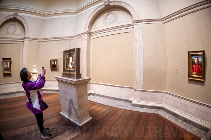 Museum visitor photographing a framed portrait in a curved gallery room lined with paintings