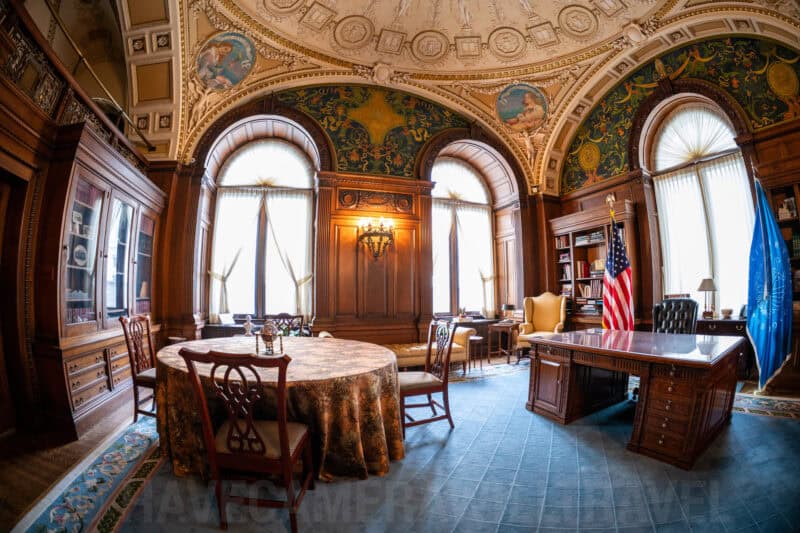 Ornately decorated study with arched windows, a round table, and an American flag beside a wooden desk