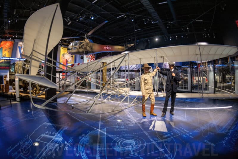 Wright brothers-style biplane replica with mannequin figures displayed in an aviation museum hall