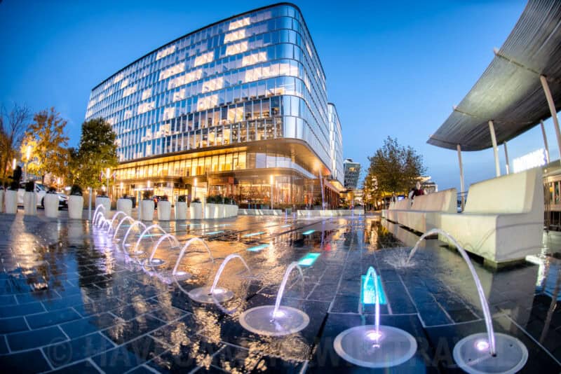 Fisheye view of a curved glass office building lit up at dusk beside a row of illuminated fountain jets