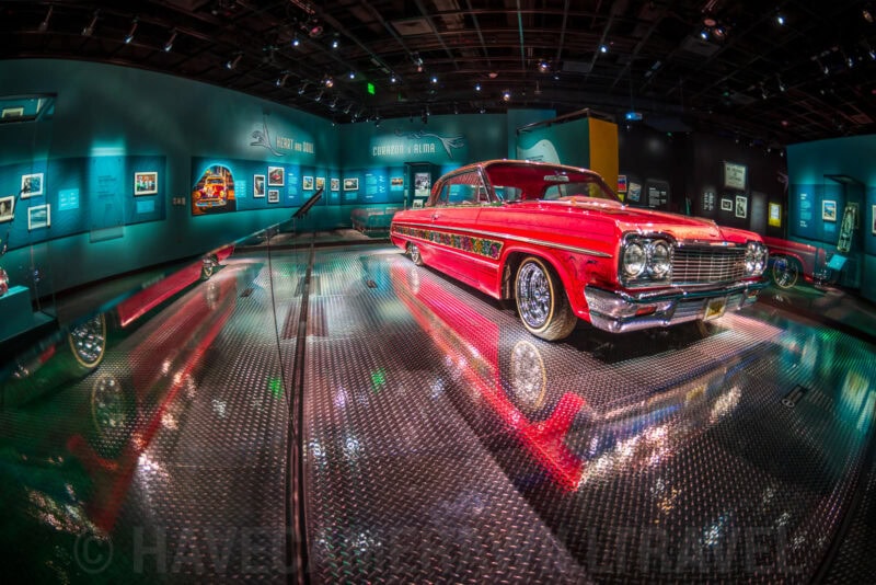 Fisheye view of a red lowrider Chevrolet Impala on display under a museum spotlight with mirrored floor