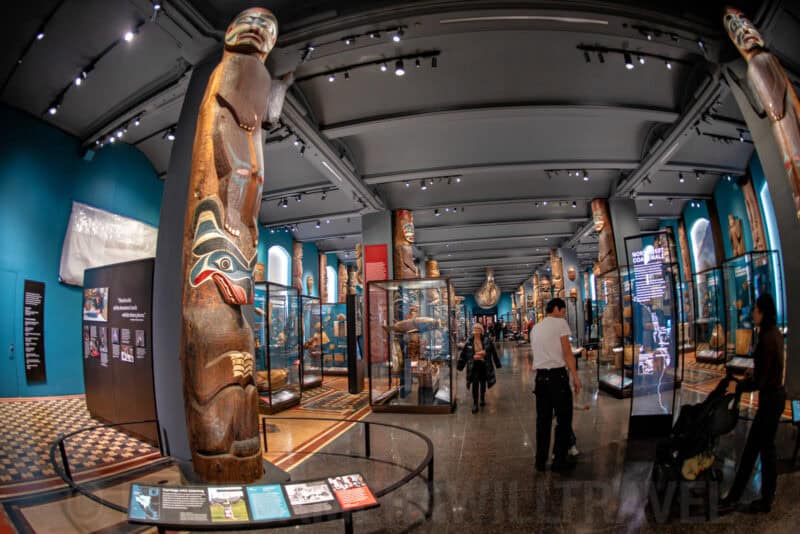 Museum hall lined with tall carved totem poles and glass display cases of Native artifacts