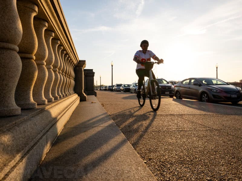 Cyclist riding past a stone balustrade with parked cars along a sunlit bridge
