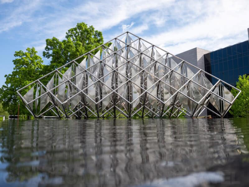 Angular mirrored steel pyramid sculpture reflected in a pool beside a glass building