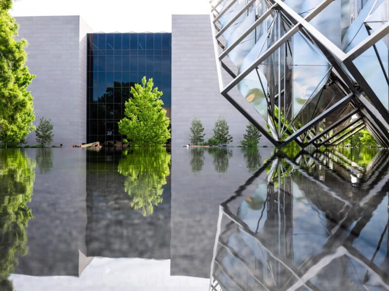 Modern glass pavilion with angular mirrored sculpture reflected in a still reflecting pool