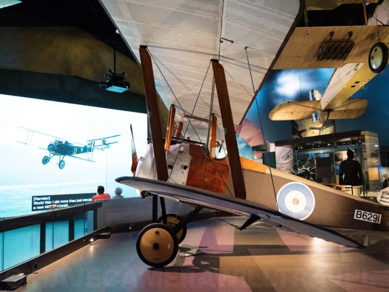 Vintage biplane displayed beneath a large video screen inside an aviation exhibit hall