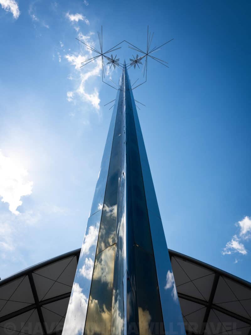 Tall mirrored steel spire sculpture rising against blue sky beside a curved pavilion roof
