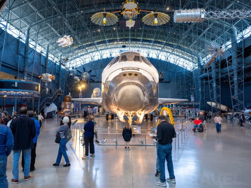 Visitors gathered around a space shuttle displayed nose-forward in a museum hangar